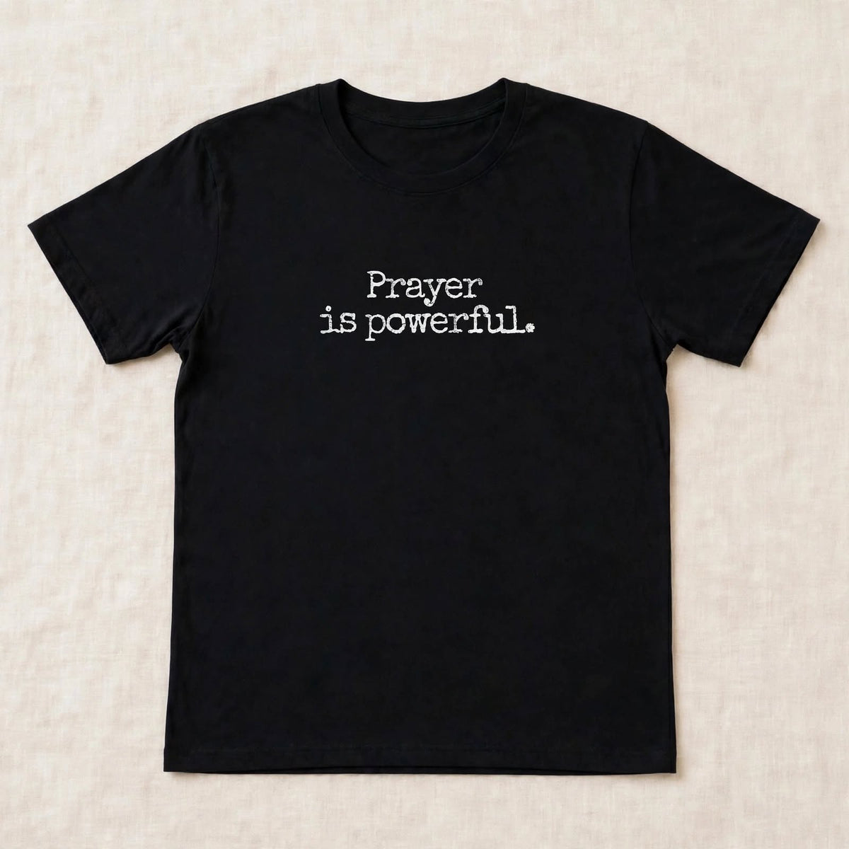 Black t-shirt flat lay on beige linen background with small centered white distressed typewriter text Prayer is powerful dot exact font match