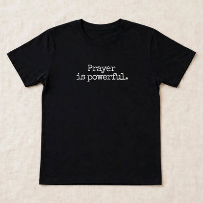 Black t-shirt flat lay on beige linen background with small centered white distressed typewriter text Prayer is powerful dot exact font match