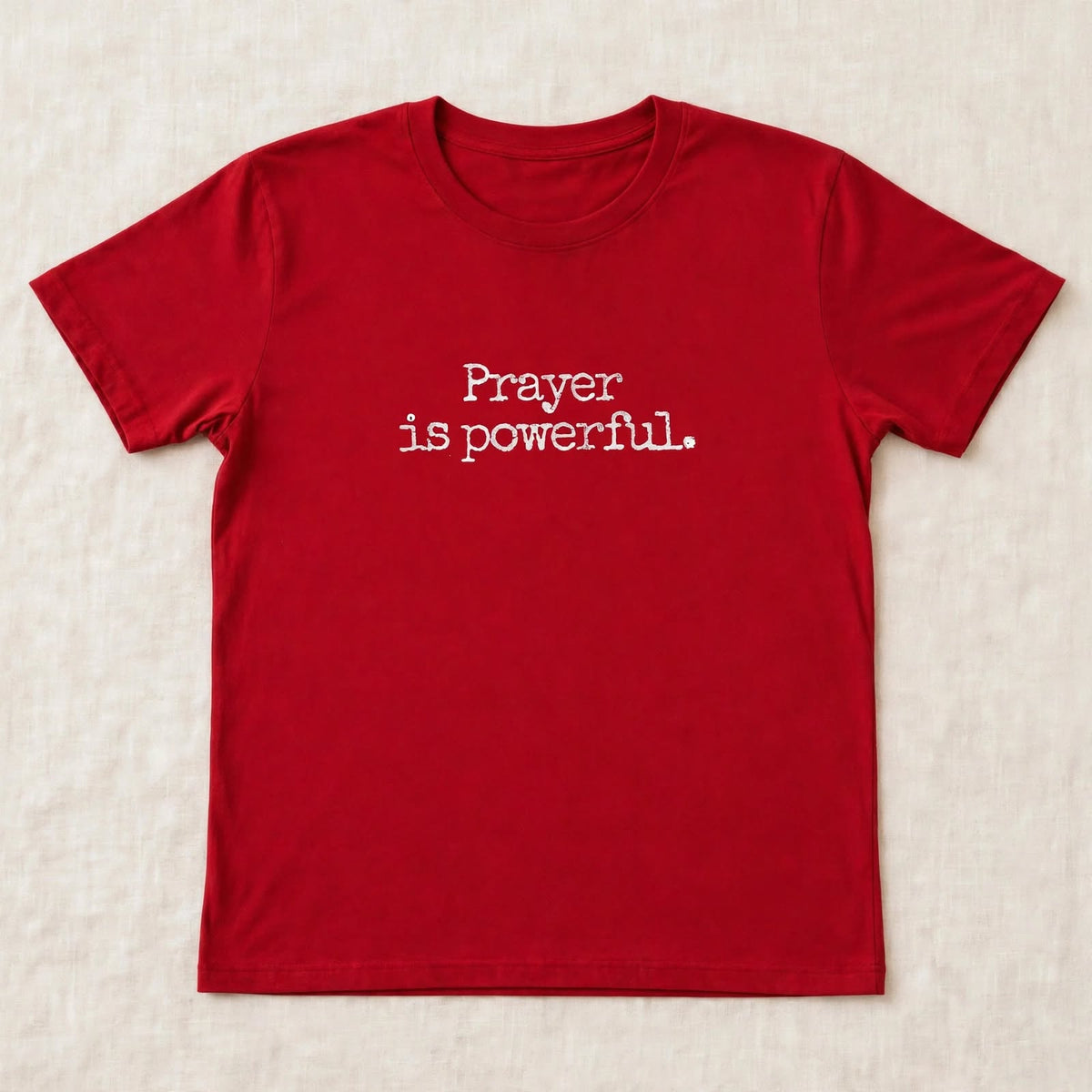 Red t-shirt flat lay on beige linen background with small centered white distressed typewriter text Prayer is powerful dot exact font match